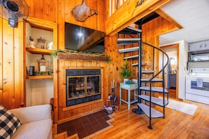 Ein Holzinnenraum mit Kamin, einem darüber montierten Fernseher, einer Wendeltreppe und einer Küchenzeile.