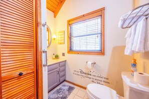 Uma casa de banho com porta de madeira, janela com persianas, sanita, barra de toalhas e um autocolante na parede com uma frase motivacional.