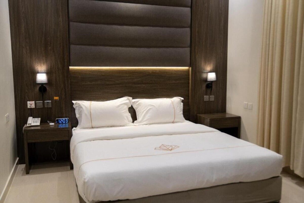 A hotel room with a large bed, two bedside tables, a wall clock, and a lighted wall panel.