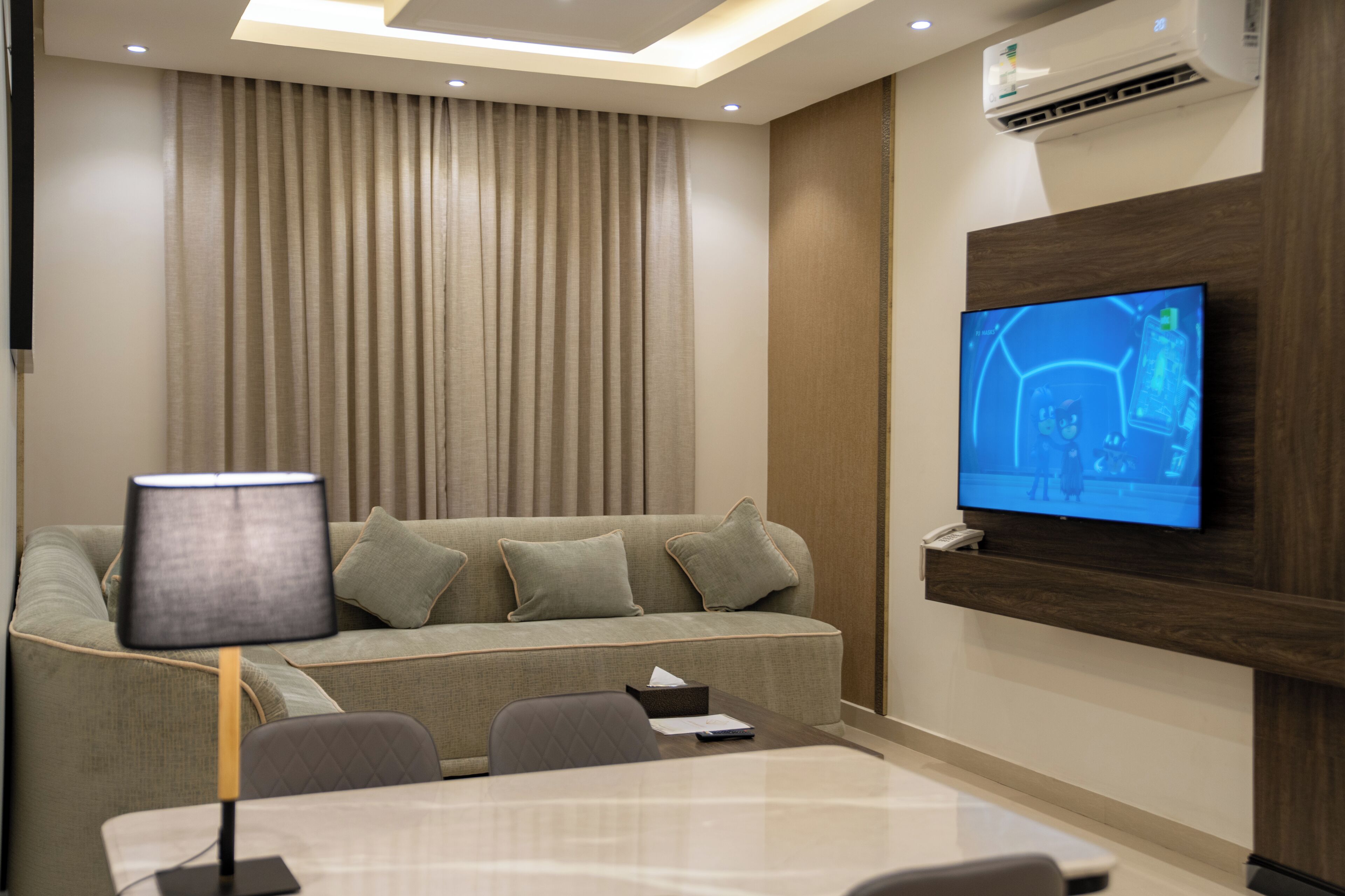 A modern hotel room with a sofa, a table, a lamp, a TV mounted on the wall, and an air conditioning unit.
