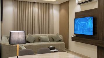 A modern hotel room with a sofa, a table, a lamp, a TV mounted on the wall, and an air conditioning unit.
