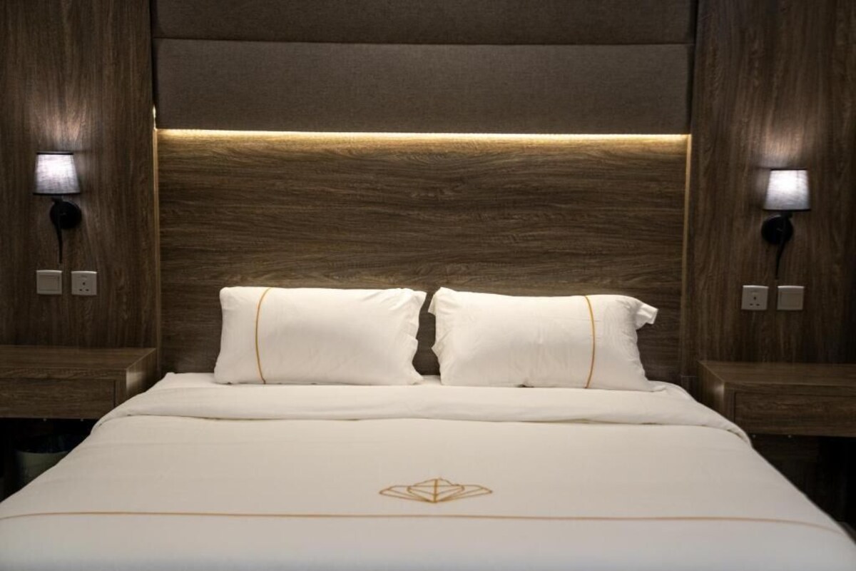 A hotel room with a wooden headboard, two bedside lamps, a bed with white bedding, and a small logo on the bedspread.