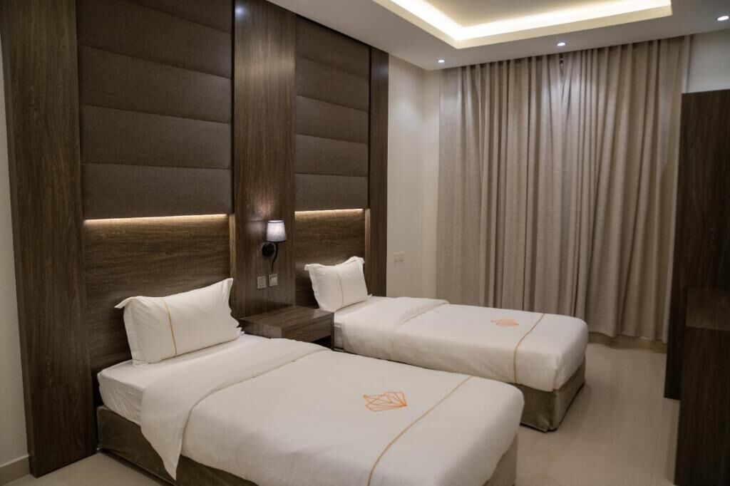 Una habitación de hotel con dos camas, cabecero de madera, mesita de noche con lámpara y un ventanal con cortinas.