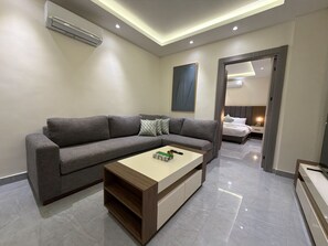 A modern living room with a grey sofa, a wooden coffee table, and a bed in the background. - 102 JRE Apartment With Stunning Boulevard Veiw (Amman)