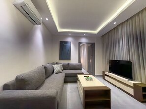 A modern living room with a sectional sofa, a flat-screen TV, and recessed ceiling lighting. - 102 JRE Apartment With Stunning Boulevard Veiw (Amman)