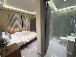 Moderni hotellihuone, jossa on sänky, yöpöytä ja lasiseinällä erotettu suihkutila.