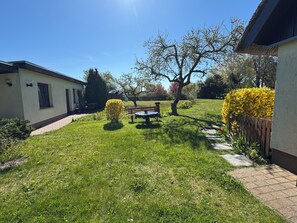Een verzorgde tuin met een patio tafel en stoelen, omringd door netjes gesnoeide hagen en diverse bomen.
