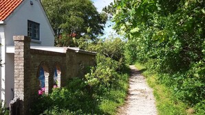 A narrow gravel path leading to a white building with a red-tiled roof, surrounded by lush greenery and trees. - This studio is located 300m from the beach and the sea. (Petten)