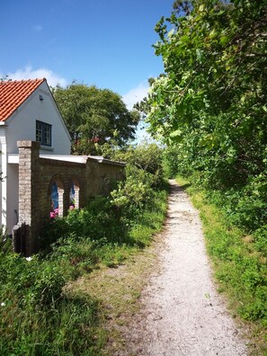 Ein schmaler Kiesweg führt zu einem weißen Gebäude mit roten Ziegeldach, umgeben von üppigem Grün und Bäumen.