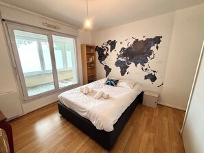 A bedroom with a world map wall, a bed with white linens, a wooden floor, and a sliding glass door to the outside. - Le millésime Vesle | Hypercentre | 6 couchages (Reims)