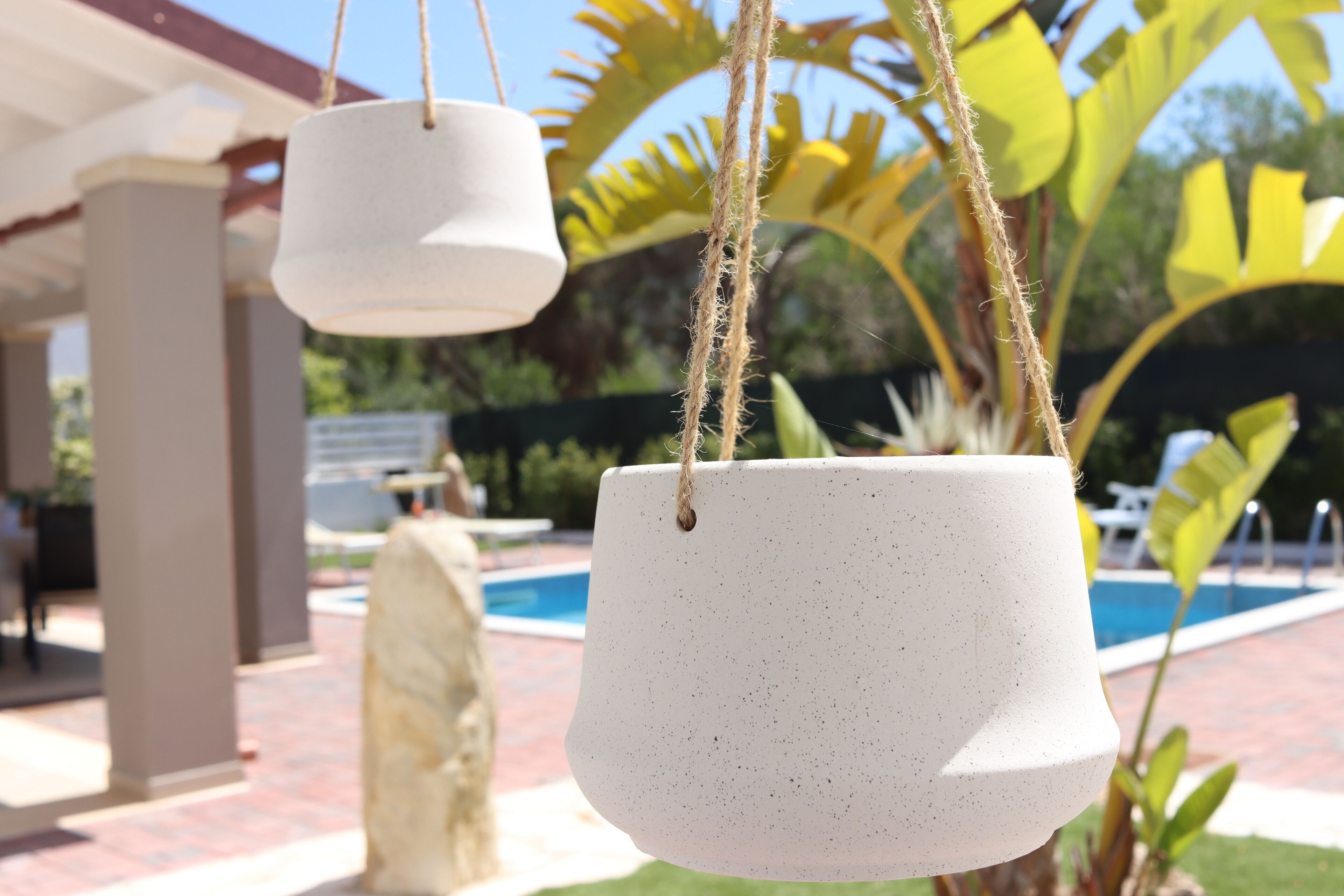Two hanging planters with rope, a pool, and a stone sculpture in the background.