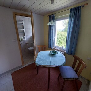 A room with a dining table, chairs, a window with curtains, a radiator, and a door leading to a bathroom. - Bungalow in Saxon Switzerland (Sebnitz)