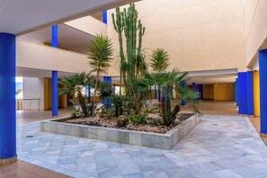 Ein geräumiger Innenhof mit einem zentralen Garten, der eine Kaktee und Palmen zeigt, umgeben von blauen Säulen und einem gefliesten Boden.
