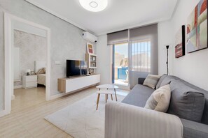 Ein modernes Wohnzimmer mit einer grauen Couch, einem Flachbildfernseher, einem kleinen weißen Tisch und einem Bücherregal.