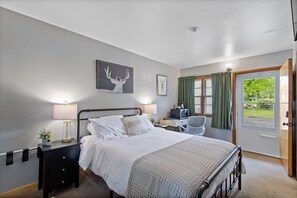 A bedroom with a bed, bedside table, lamp, chair, desk, and a view of the outdoors.