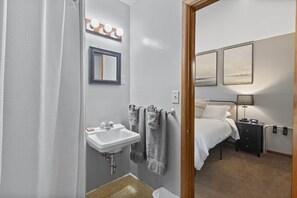 A bathroom with a sink, mirror, towel rack, and a bed visible through an open door.
