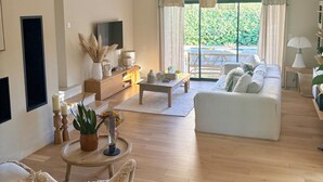Ein geräumiges Wohnzimmer mit einer Couch, einem Couchtisch und einem Fernseher. Es gibt ein Fenster mit Vorhängen, eine Pflanze und eine Lampe.