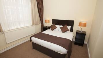 A neatly made bed with a brown headboard, two bedside lamps, and a window with sheer curtains.