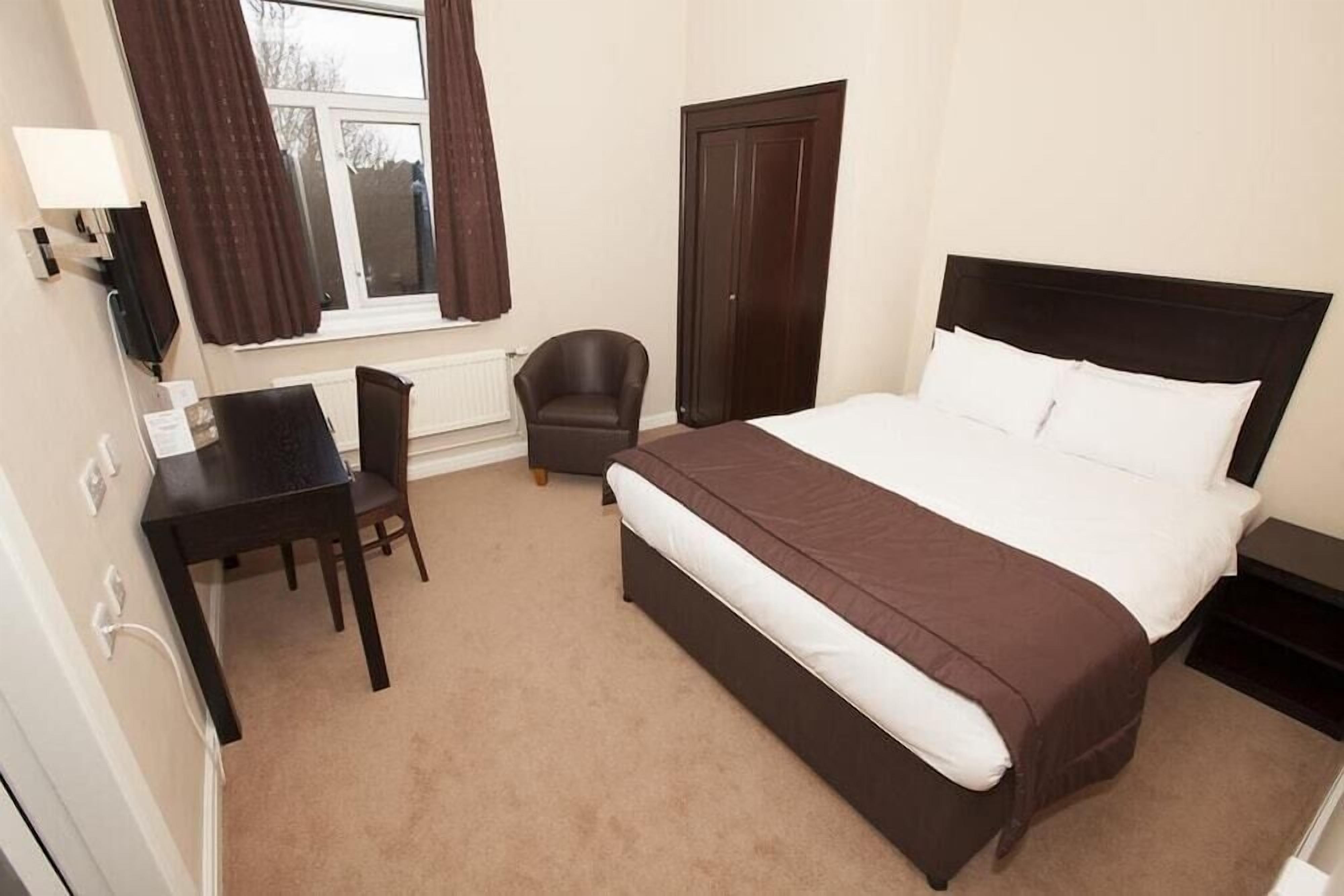 Ein Hotelzimmer mit einem Bett, einem Schreibtisch, einem Stuhl, einem Kleiderschrank und einem fensterbepflanzten Fenster.