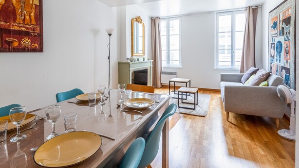A dining area with a table set for four, a fireplace, and a living room with a sofa and a wall art piece. - L'Heure Dorée - Duplex under the Grosse Horloge (La Rochelle)