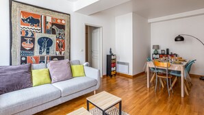 A living room with a sofa, a small table, a dining area with chairs, a wall with a colorful tapestry, and a door leading to another room. - L'Heure Dorée - Duplex under the Grosse Horloge (La Rochelle)
