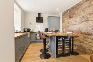 A modern kitchen with grey cabinets, a wooden countertop, a central island with stools, a stone wall, and a built-in wine cooler. - Harbour Cottage Seahouses (Seahouses)