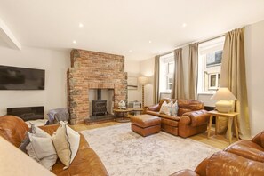 A cozy living room with a brick fireplace, leather sofas, a flat-screen TV, and a large window with curtains. - Harbour Cottage Seahouses (Seahouses)