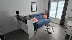 Ein kompakter Wohnraum mit Küchenzeile, einer Couch mit Kissen, einem kleinen runden Tisch und einem an der Wand befestigten Fernseher.