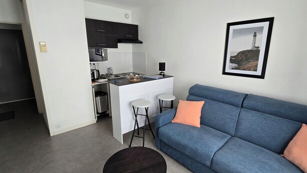 Ein kleiner Wohnraum mit einer Küchenzeile, einem blauen Sofa mit orangefarbenen Kissen und einem gerahmten Bild an der Wand.