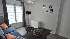 Ein Wohnzimmer mit einer Couch, einem Couchtisch mit einer Schale Snacks, einem Fernseher an der Wand und einem Fenster mit Jalousien.
