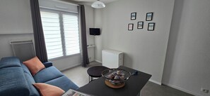 Ein Wohnzimmer mit einer Couch, einem Couchtisch mit einer Schale Snacks, einem Fernseher an der Wand und einem Fenster mit Jalousien.