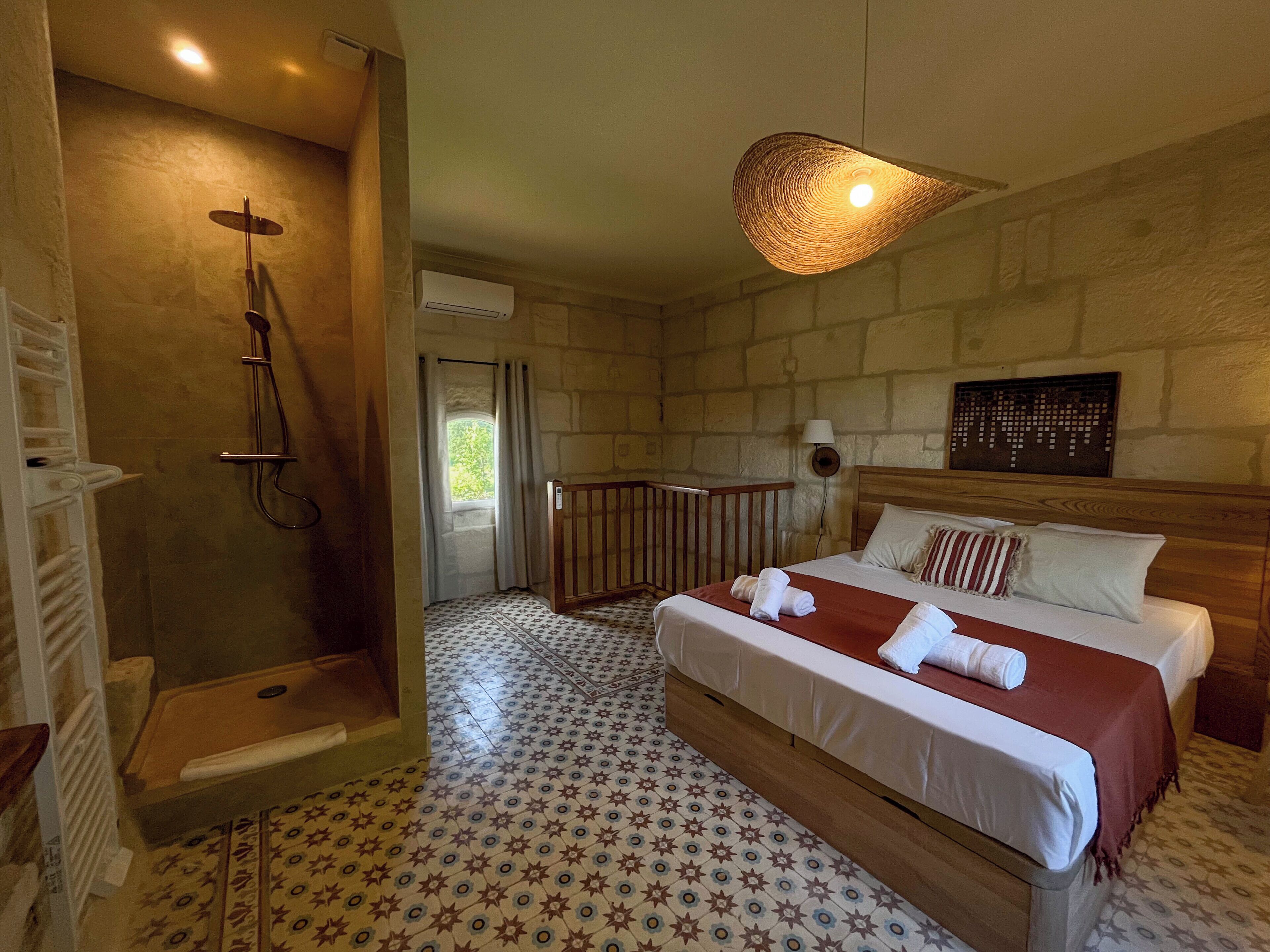 Une chambre d’hôtel avec une douche, un lit recouvert d’une couverture rouge, un sol à motifs et un mur en pierre.