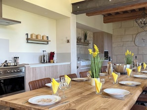 A dining area with a long wooden table set for a meal, kitchen appliances, and a clock on the wall.