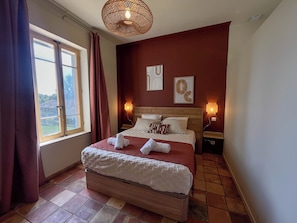 A bedroom with a large bed, two bedside tables, a window with curtains, a wall-mounted lamp, and artwork.