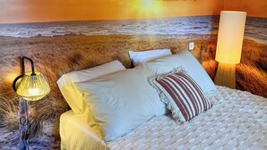 Ein Bett mit weißen Laken und einem gestreiften Kopfkissen, eine Lampe und ein Wandbild mit einem Sonnenuntergang über dem Meer.