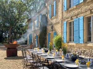 Un área de comedor al aire libre con una larga mesa dispuesta para una comida, rodeada de edificios de piedra con contraventanas azules.