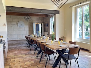 A dining area with a long table set for six, surrounded by chairs, with a stone wall and a large window in the background. - Mas Dou Rose, farmhouse with swimming pool near Alpilles (Arles)