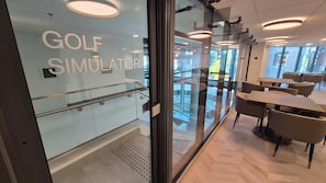 Un espai d'oficina modern amb una paret de vidre que diu 'GOLF SIMULATOR', una escala i una zona de menjador amb taules i cadires.