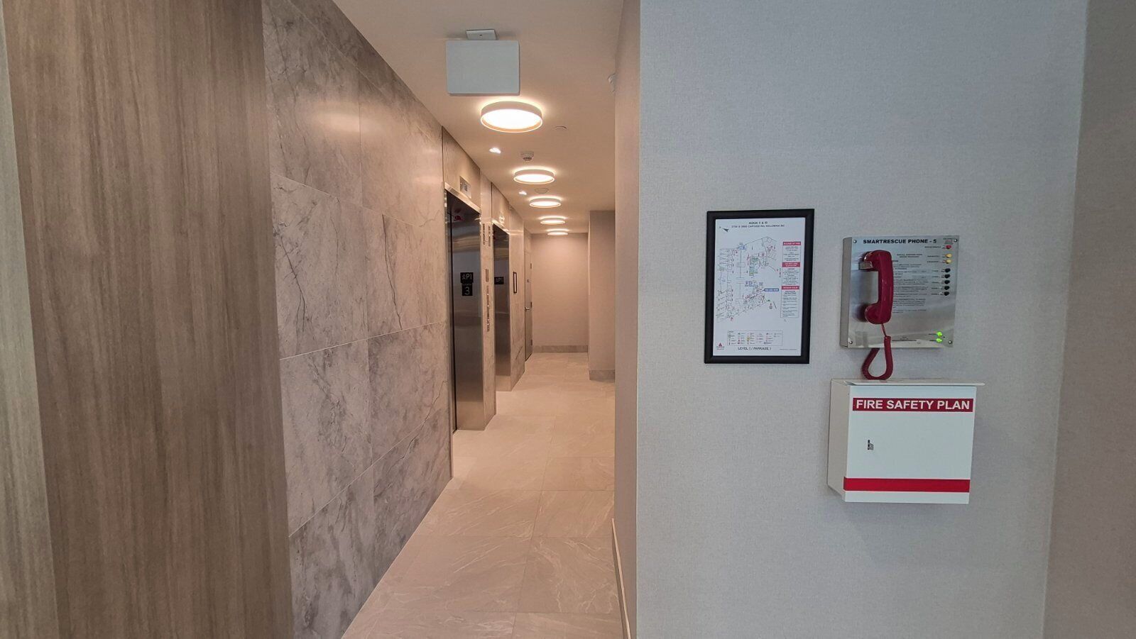 A hallway with marble walls, a fire safety plan sign, a smoke detector, and a fire alarm pull station.