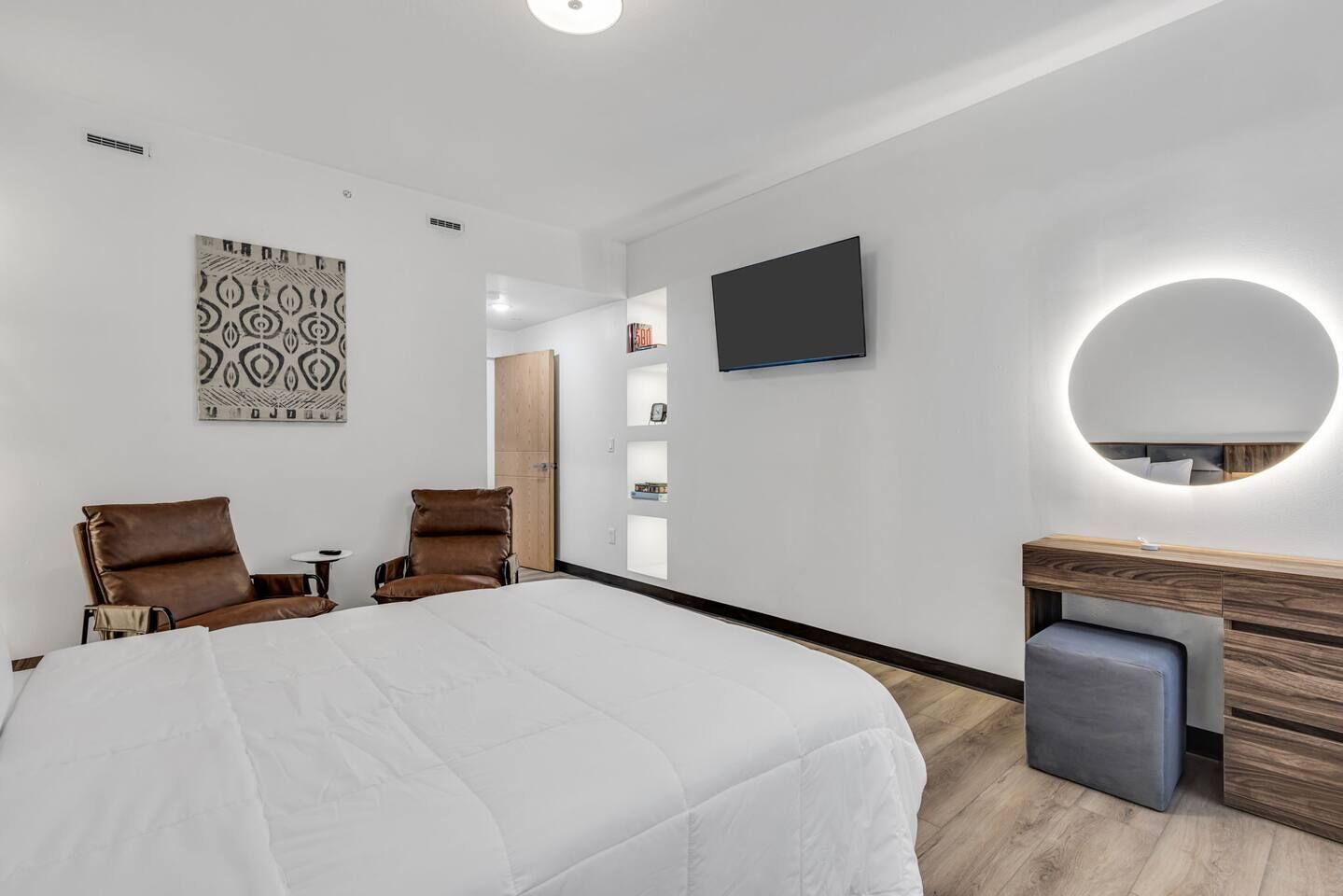 Una habitación de hotel moderna con una cama grande, dos sillones de cuero, un tocador de madera, un televisor y un adorno mural con estampado.