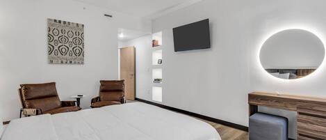 Una habitación de hotel moderna con una cama grande, dos sillones de cuero, un tocador de madera, un televisor y un adorno mural con estampado.