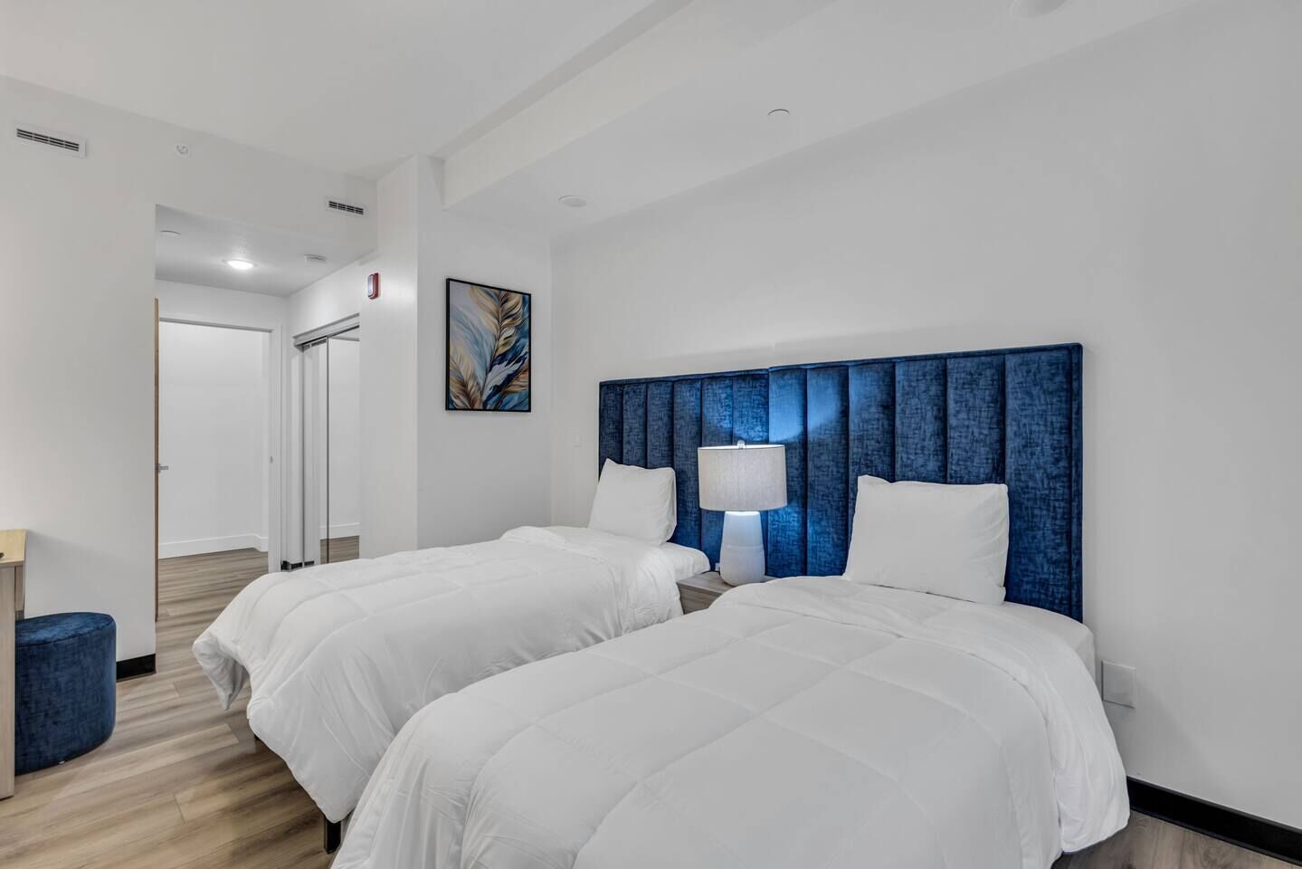 A hotel room with two beds, a blue headboard, a painting on the wall, and a lamp on the nightstand.