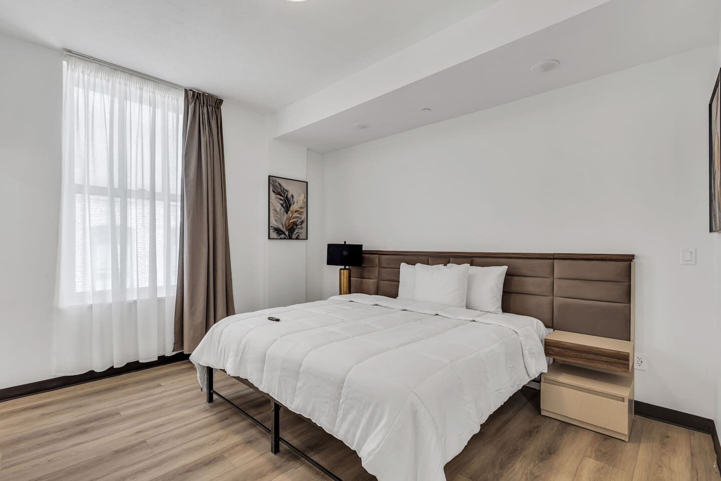 A modern hotel room with a large bed, a bedside table, a lamp, and a window with curtains.