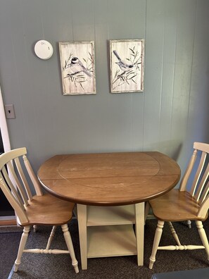 A round wooden table with two chairs, a wall-mounted smoke detector, and two framed paintings of birds.