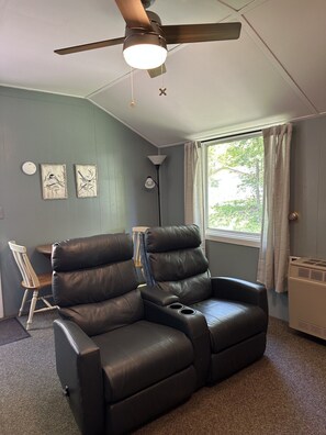 A room with a ceiling fan, two wall-mounted bird paintings, a window with curtains, a black leather recliner, a wooden chair, and a floor lamp.