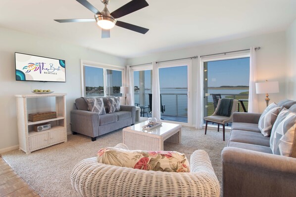 A spacious living room with a grey sofa, a wicker chair, a coffee table, and a television displaying 'The Best Rentals' logo. - Sunset Corner ~ Surrounding river views (Folly Beach)