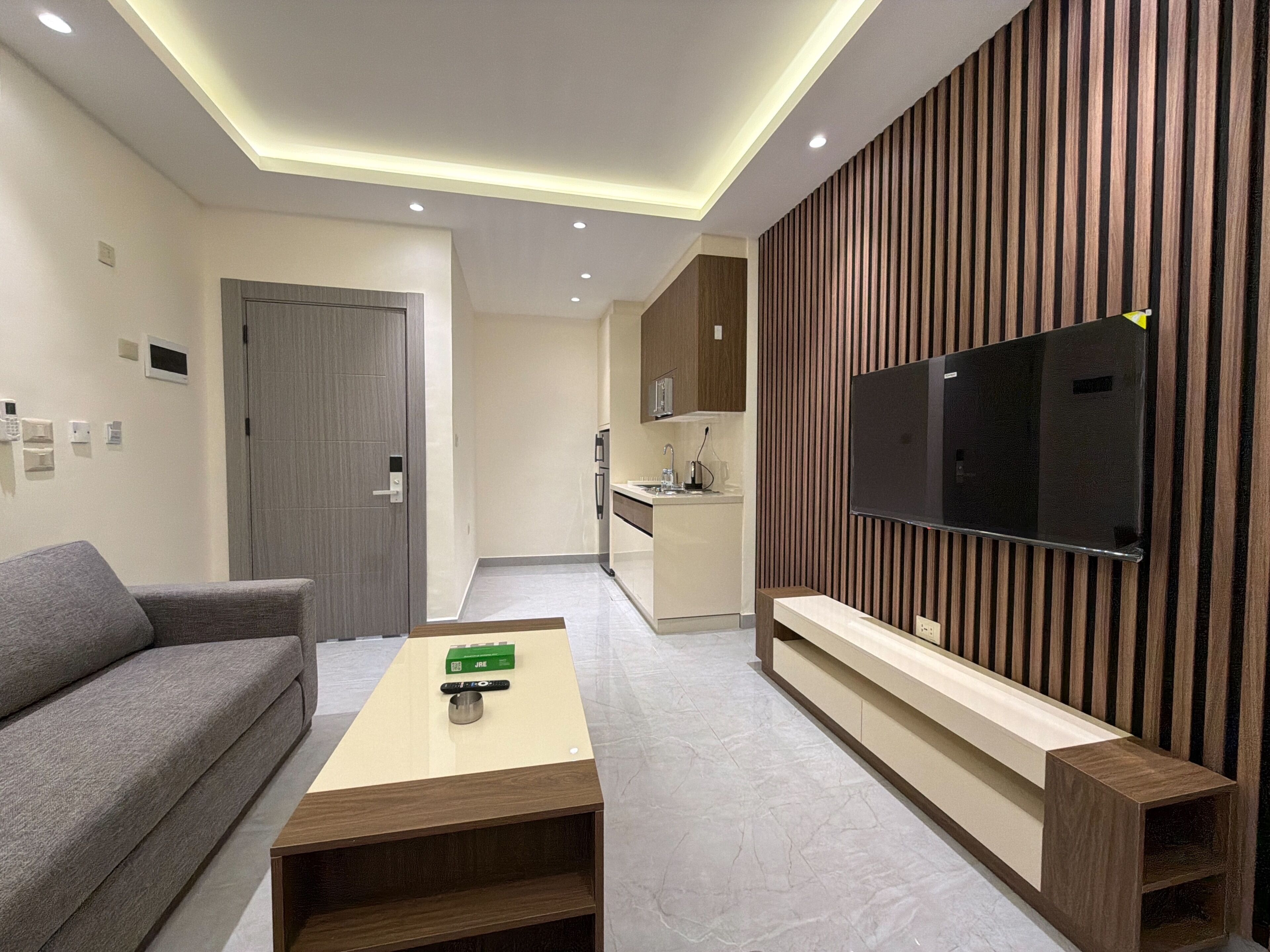 A modern living room with a sofa, a coffee table, a TV mounted on a wooden panel wall, a kitchen area in the background, and recessed ceiling lighting.