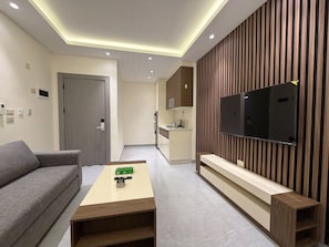 A modern living room with a sofa, a coffee table, a TV mounted on a wooden panel wall, a kitchen area in the background, and recessed ceiling lighting. - 103 JRE Comfortable and Accessible Retreat (Amman)
