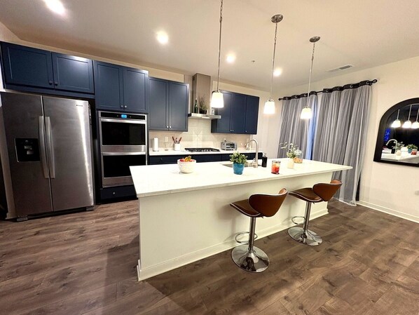 A modern kitchen with a central island, bar stools, stainless steel appliances, and pendant lighting.