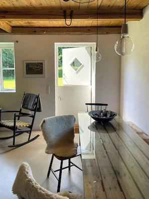 Ein Zimmer mit Holzboden, einer weißen Tür mit kleinem Fenster, einem Schaukelstuhl, einem gemütlichen Sessel, einem Esstisch mit einer Schüssel und einer Hängelampe.